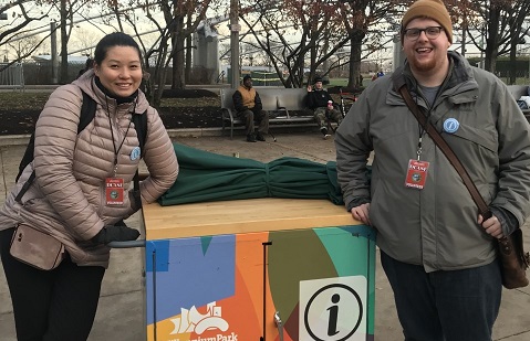 Two smiling volunteers in coats stand with the Millennium Park information cart outside in Millennium Park during a volunteer shift