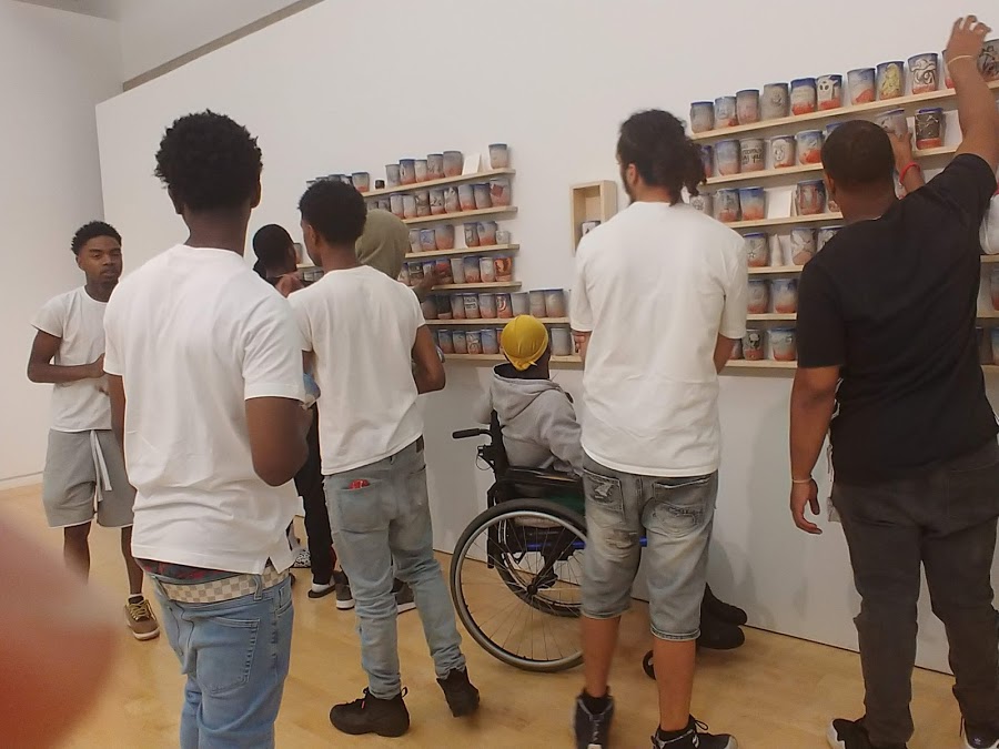 a group of teens and their chaperone explore the art in an exhibition at the Chicago Cultural Center - they are all wearing white shirts except one and one teen uses a wheelchair