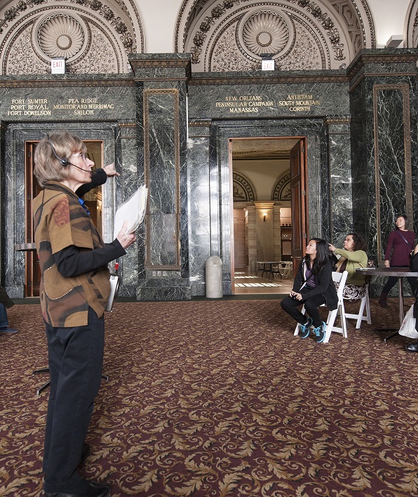 Volunteer Judy is wearing a headset and pointing to the walls in Grand Army Hall as she leads a free private tour for a preregistered group